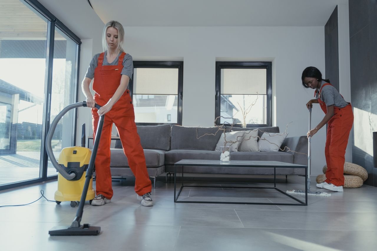 Services Two women in red overalls vacuuming and sweeping a modern living room.