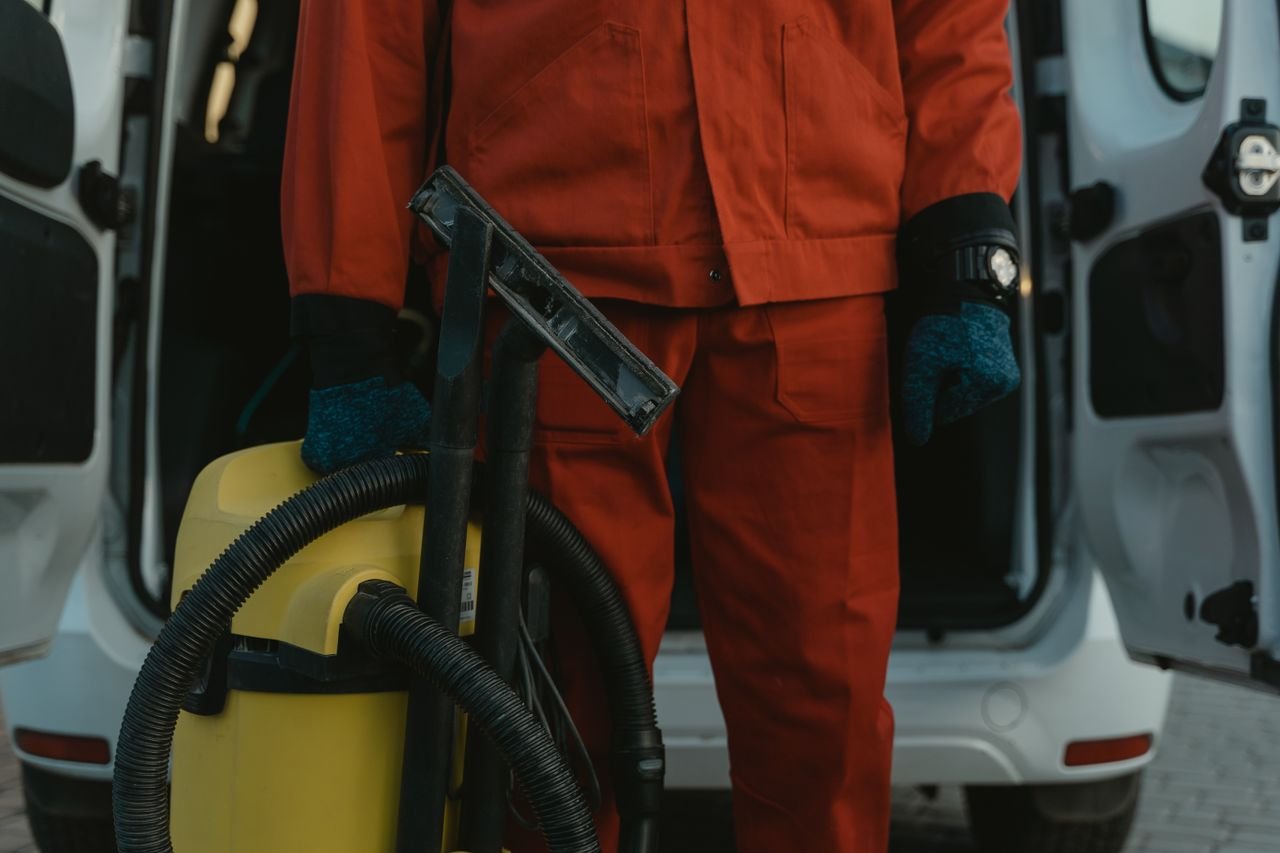 Services A professional cleaner in a red uniform holding a vacuum near a van, ready for service.